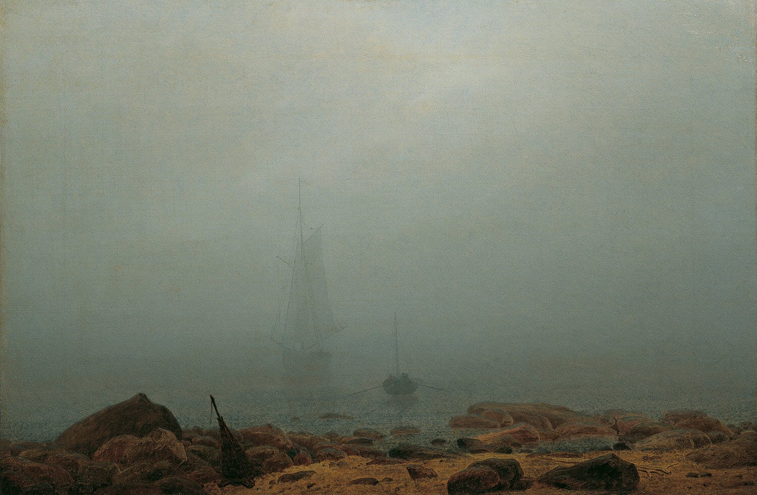 Das Gemälde „Meeresstrand im Nebel“ von Caspar David Friedrich zeigt eine ruhige Küstenlandschaft im dichten Nebel. Im Vordergrund sind einzelne Felsen und Treibholz sichtbar, während der Horizont im diffusen Licht verschwimmt. Die Szene strahlt Stille und Melancholie aus.